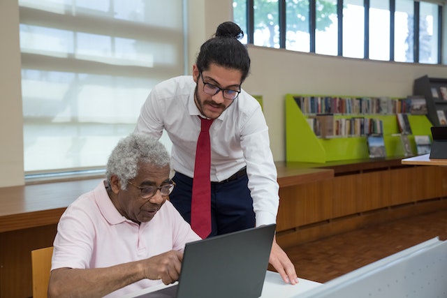 Picture of a young man assisting an older man on a desktop in a public library.