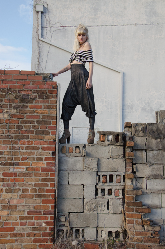 a  portiat of a young woman in flowy pants, a small striped shirt, and lace up boot high heals standing ontop of a broken brick wall