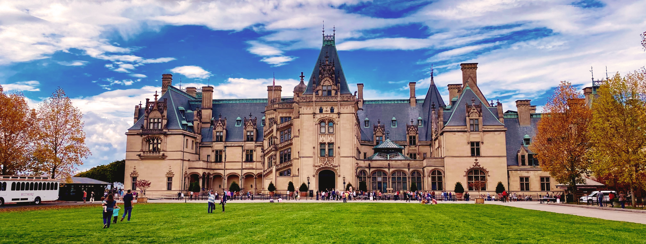 The front of the Biltmore house with people outside and on the lawn on a partially cloudy fall afternoon.